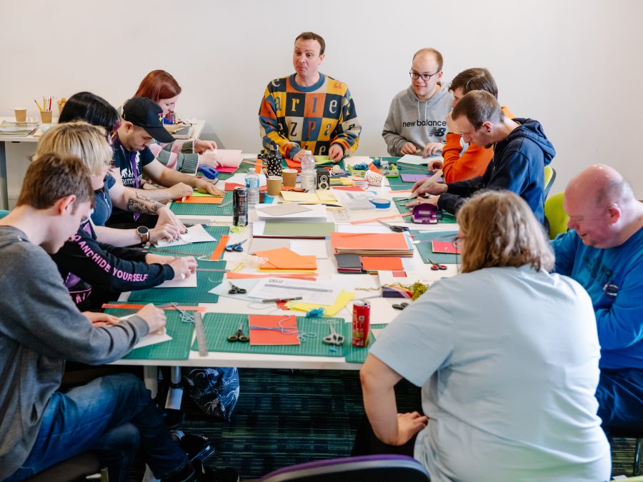 A group of people sit around a large table covered with colourful paper, pens, and craft materials during a creative workshop. They appear focused on their projects, with a facilitator seated at the far end guiding the activity in a bright, plain room.