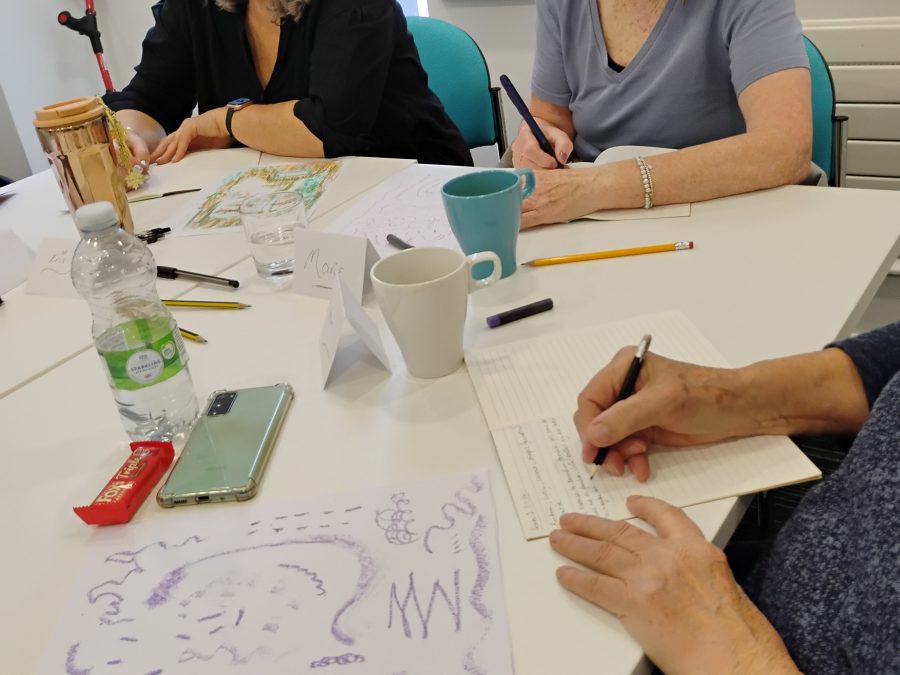 This image shows three individuals sitting at a table engaged in an activity that involves writing and drawing. The individuals' faces are blurred for privacy. On the table, there are various items including a water bottle, a smartphone, a red snack bar, a pair of glasses, a few cups, and some papers. One of the papers has a drawing made with purple lines and shapes. Another individual is writing in a notebook with a black pen. There are also name tags on the table.
