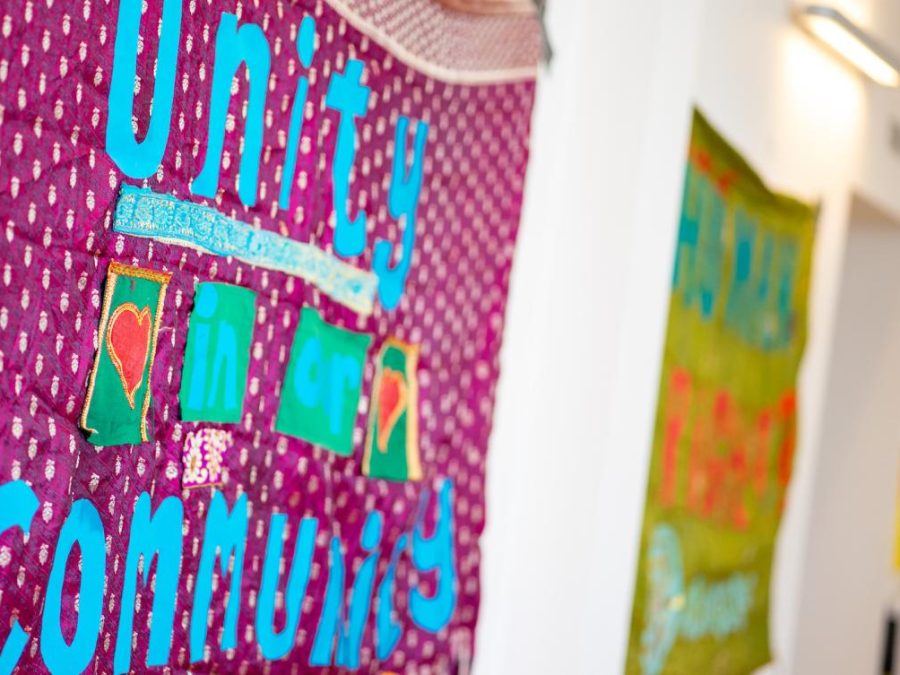 A handmade fabric banner with a deep maroon background and small white patterns hangs on a wall. Bright blue letters spell out “Unity in our Community,” with the words “in” and “our” displayed on green patches bordered in gold. Two additional green patches feature red hearts outlined in gold. A light blue decorative strip runs horizontally across the banner. In the background, another colorful fabric banner is visible but out of focus. Image credit. Banners by Jane Thakoordin (shown at the CHWA National conference 2023)