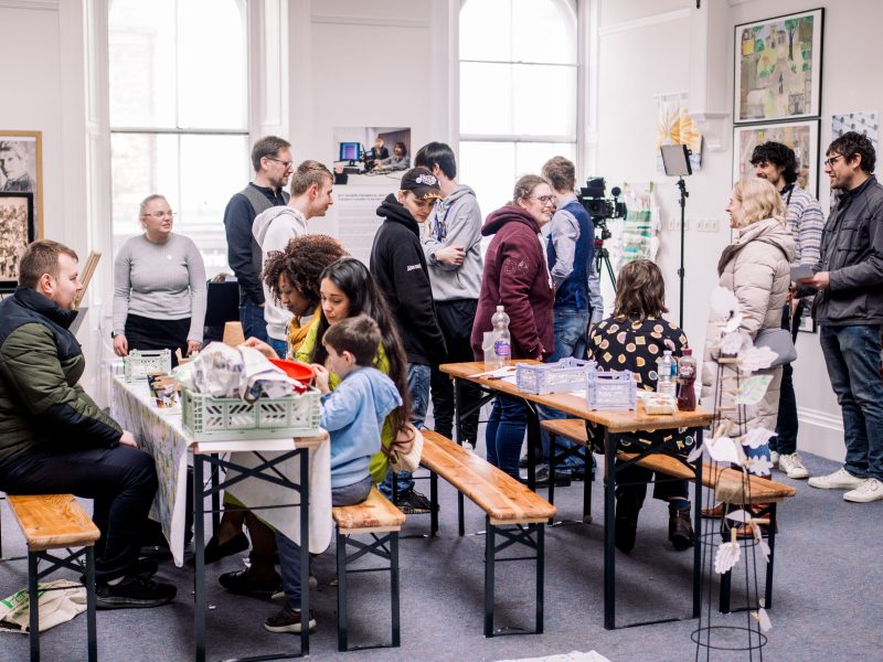 A lively community workshop taking place in a bright room with large windows. Several people are seated at long wooden tables covered with craft materials, fabric, and baskets, while others stand nearby chatting and observing. The group includes adults and children engaged in creative activities. Posters, artwork, and photographs decorate the walls, and a camera setup suggests documentation or filming.